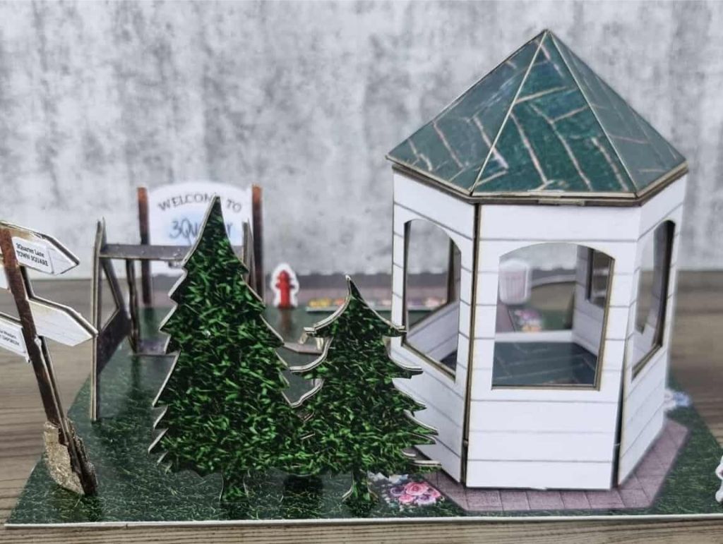 Miniature park scene with white gazebo, pine trees, swing set, and welcome signage for 3Quarter Lane, set on a grass-textured base.