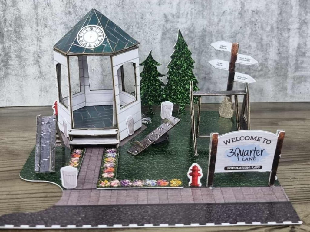 Miniature town square scene featuring a white gazebo with green roof, clock tower, directional signpost, and welcome sign for 3Quarter Lane.