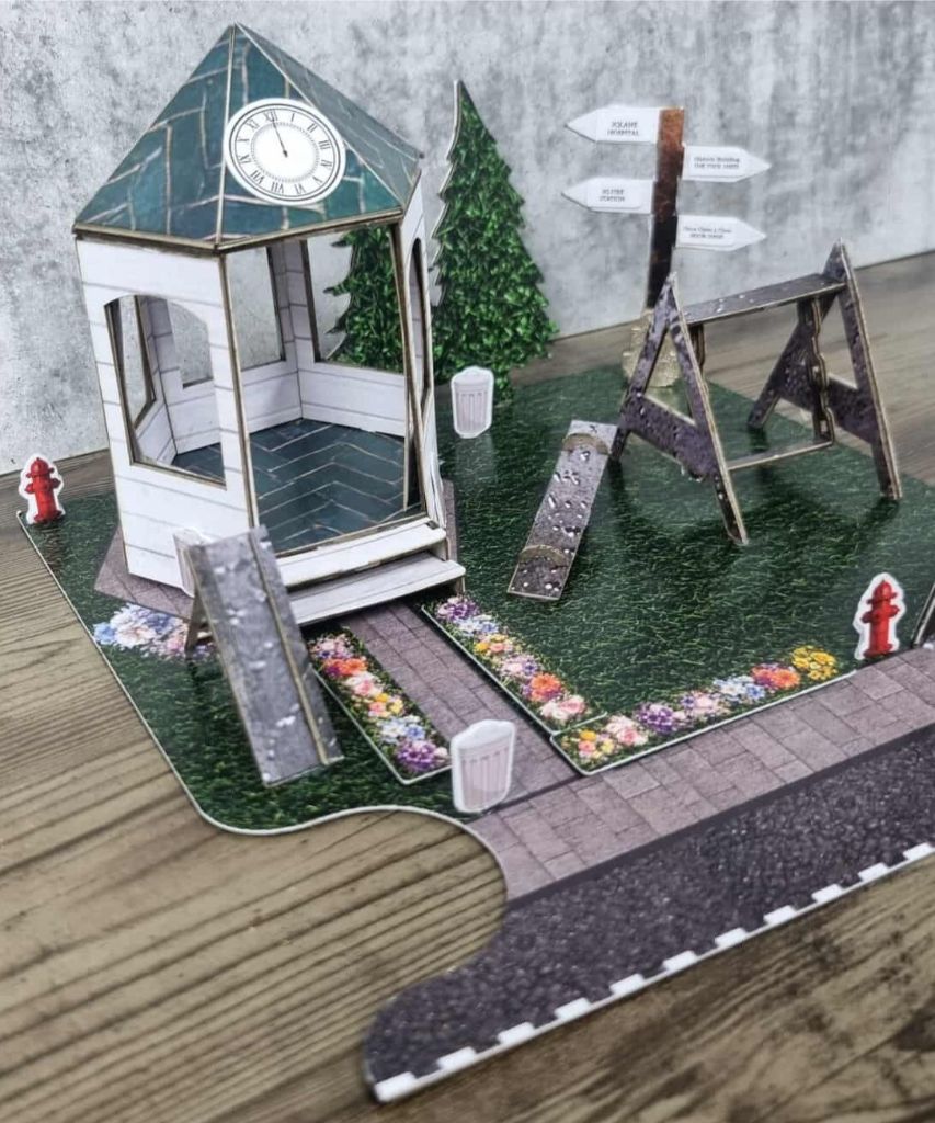 Detailed paper diorama of a community park with gazebo, swing set, flower-lined path, fire hydrants, and directional signs.