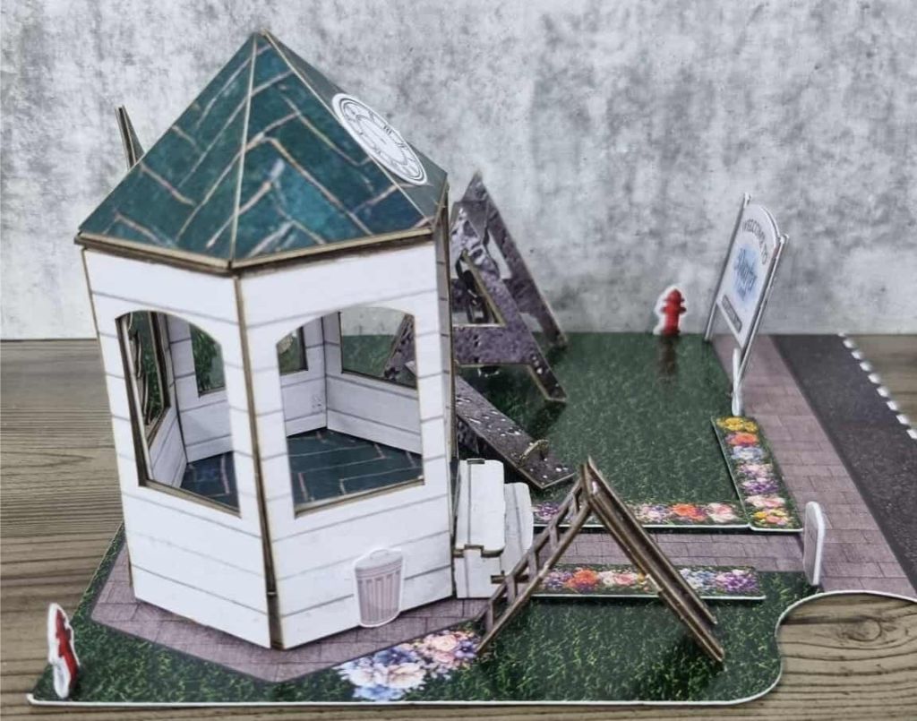 Miniature town square with white gazebo, green roof, flower beds, and welcome signage in a realistic park setting.