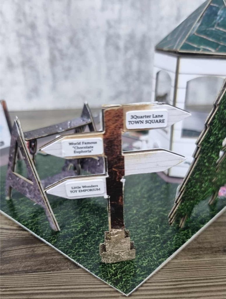 Miniature town square scene with signpost pointing to 3Quarter Lane Town Square, Chocolate Euphoria, and Little Wonders Toy Emporium.